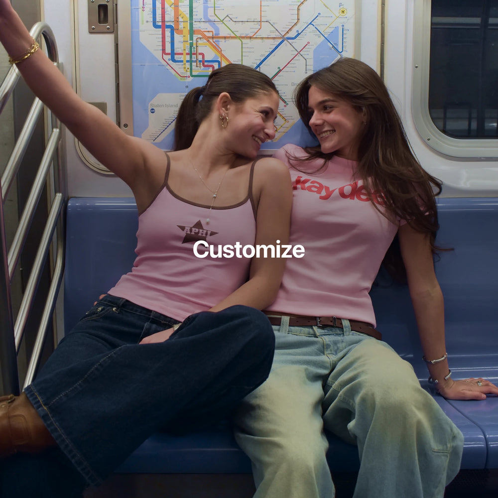 Two girls sitting on a subway bench wearing pink sorority merch with visible text.